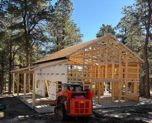 A wooden-framed house under construction in a wooded area, with a red compact construction vehicle parked in front.