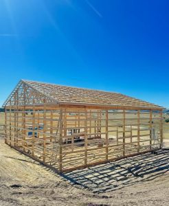 A wooden frame of a rectangular building under construction stands on a dirt lot under a clear blue sky.