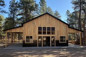 A wooden building with a peaked roof and large windows stands in a forested area, surrounded by tall pine trees under a clear sky.