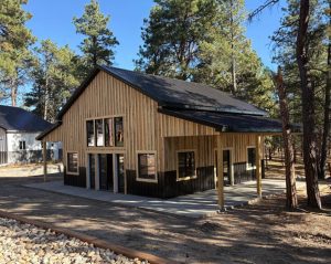 A modern wood and metal building with large windows, set among tall pine trees on a concrete foundation, under a clear blue sky.