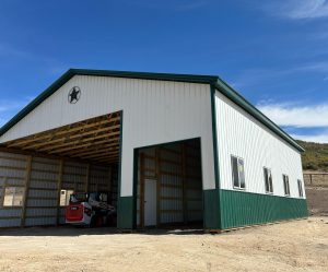 A large metal building with a green base and white upper walls, partially open, houses a skid steer loader under a clear sky.