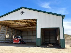 A white and green metal barn with a large open doorway and a red skid steer parked inside; a star decorates the front gable.