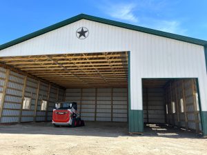 A red and white skid steer loader parked inside a large open-sided metal barn with a black star decoration on the front.