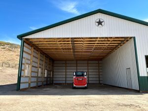 A red tractor is parked inside a large open-sided metal barn with a green roof and a star emblem on the front.