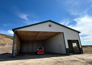 A large metal barn with a green trim and a star above the door, housing a small red and white utility vehicle, sits on a dirt lot under a partly cloudy sky.