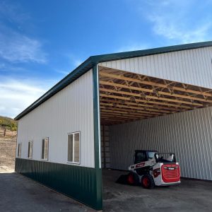 A white and green metal building with open front, showing exposed roof beams and a parked Bobcat skid steer loader inside.