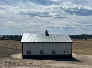 A metal-roofed building with four windows sits on a dirt lot, surrounded by open fields and a partly cloudy sky.