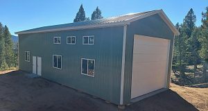 A large green metal building with multiple windows, a white door, and a tall garage door, surrounded by trees and dirt ground under a clear blue sky.
