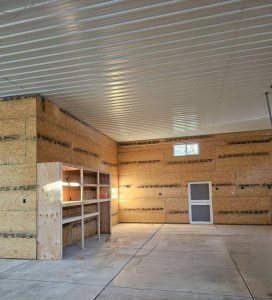 A spacious, unfinished garage with wood-paneled walls, concrete floor, a small window, a door, and a wooden shelving unit in one corner.