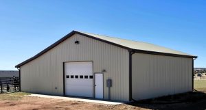 A light-colored metal storage building with a gable roof, a large overhead garage door, a single entry door, and an electrical box on the exterior wall.