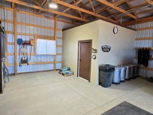 Interior of a tidy utility or tack room with wooden beams, metal walls, hanging tools, trash cans, grain bags on a pallet, and a clock above a door on a tan partition.
