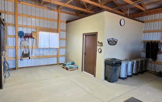 Interior of a tidy utility or tack room with wooden beams, metal walls, hanging tools, trash cans, grain bags on a pallet, and a clock above a door on a tan partition.