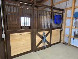 A clean horse stall with wooden lower panels, metal bars on top, a blue fan mounted on the wall, and ropes hanging on the stall door.