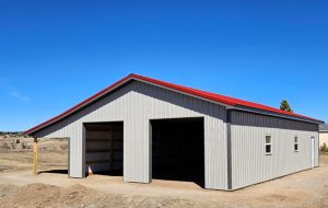 A large, light gray metal garage with two open doors and a red roof stands on a dirt lot under a clear blue sky.
