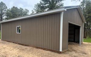 A brown metal-sided garage or shed with a partially open overhead door and one small window, situated on a dirt patch and surrounded by trees.