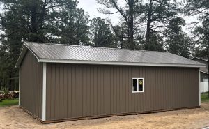 A brown metal-sided shed with a metal roof and one small window, situated on a dirt patch, surrounded by trees.