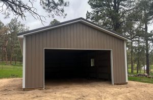 A brown metal garage with an open door stands on a dirt area, surrounded by trees and grass under a cloudy sky.