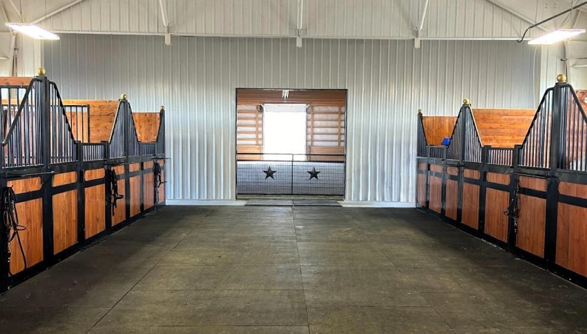 Interior of a clean, well-lit horse stable with empty stalls, black metal railings, and wooden panels—showcasing effective riding arena dust control—leading to a closed door adorned with two star decorations.