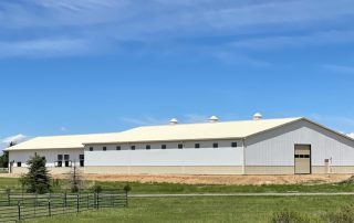 A large white industrial building with small windows and a garage door sits on a grassy lot under a blue sky with scattered clouds, ideal for use as a riding arena with dust control solutions.
