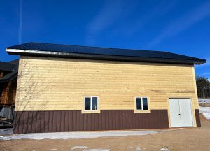 A large wooden building with a metal roof, two small square windows, and a white double door, standing on a dirt and snow-covered ground under a clear blue sky.