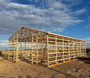 A wooden frame structure of a large building under construction stands on a dirt lot, with suburban houses and a blue sky with clouds in the background.