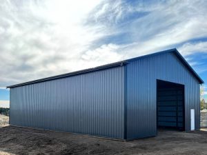 A large blue metal storage building with an open entrance stands on bare ground under a partly cloudy sky.