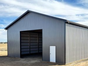 A large gray metal storage building with an open garage door and a small white entrance door, set on a dirt lot under a partly cloudy sky.
