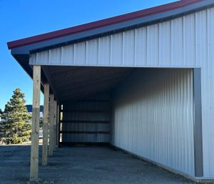A metal pole barn structure with a sloped roof, partially enclosed on three sides, stands on bare ground under a clear blue sky.