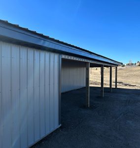 A metal building with a tin roof and open support posts stands on a dirt area under a clear blue sky.
