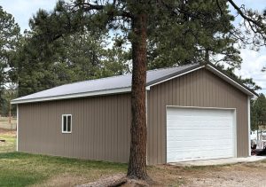 A brown metal-sided garage with a white roll-up door and a small window, situated under pine trees on a grassy lot.