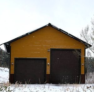 A yellow building with two large brown garage doors sits in a snowy landscape under a cloudy sky.