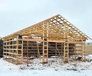 Wooden frame of a barn or shed under construction on a snowy ground, with open sides and visible trusses and beams.