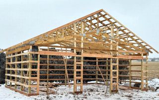 Wooden frame of a barn or shed under construction on a snowy ground, with open sides and visible trusses and beams.
