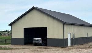 A large metal and wood garage building with a partially open door, showing the front of a pickup truck inside on a gravel driveway.