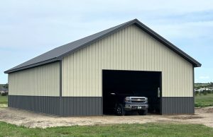 A large metal storage building with beige siding and a dark roof, partially open to reveal a parked pickup truck inside.