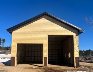 A wooden garage with two large open doorways, set on a dirt area with patches of snow and trees in the background under a clear blue sky.