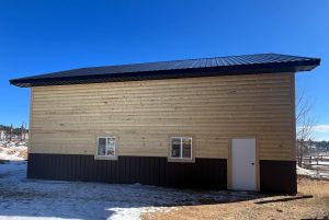 A tan and dark brown metal-sided building with a black roof, two windows, and a white door, set on snowy ground under a clear blue sky.