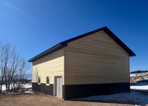 A beige and brown metal building with a peaked roof stands on bare ground, with snow patches and leafless trees nearby under a clear blue sky.