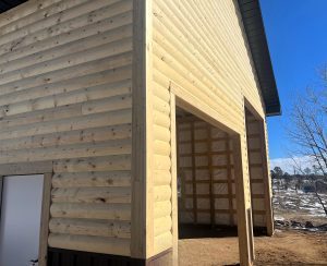 A large wooden building with log-style siding, a wide open doorway, and a small white door, with sunlight and some snow visible outside.