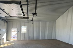 Interior of an empty metal garage with a concrete floor, a window, an open door, and an automatic garage door opener mounted on the ceiling.