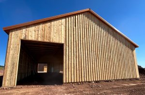 A large wooden barn with an open entrance sits on dirt ground under a clear blue sky. Sunlight casts shadows inside the unfinished structure.