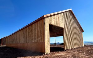 A large wooden barn structure under construction stands on bare earth, with an open entrance and clear blue sky in the background.
