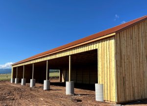 A large, unfinished wooden barn with open bays and a rust-colored metal roof stands on a dirt lot under a clear blue sky.