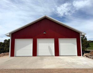 A red metal building with three closed white garage doors, standing on a concrete driveway under a partly cloudy sky.