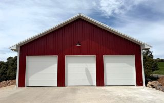 Red pole barn with three white garage doors and a concrete driveway under a partly cloudy sky.