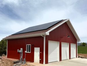 Red garage building with three white doors and rooftop solar panels, set on a concrete driveway with electrical panels mounted on the side.