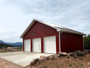 A red metal garage with three white roll-up doors stands on a concrete pad surrounded by rocks and sparse vegetation under a cloudy sky.