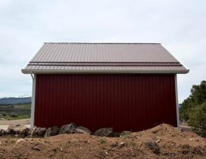 A red metal-sided barn with a silver metal roof sits on a dirt foundation, with rocks in front and trees and mountains in the background under a cloudy sky.