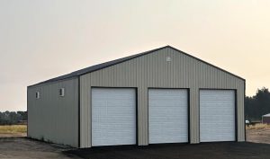 A large metal building with three white garage doors, small side windows, and a gable roof, situated on a dark paved surface in a rural area.