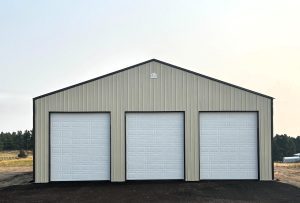 A beige metal building with three large white garage doors stands on a paved surface, with trees and grass visible in the background.